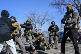 Four federal agents stand in a street holding weapons; their body language appears tense, alert, and aggressive. One agent appears to be confronting a civilian in the foreground. In the background, agents tackle a protester.