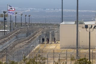 Wide shot of a detention facility with two fences topped with barbed wire surrounding it. Heat waves ripple and distort the shot. Three distant figures walk into the facility. Flags outside of the facility - California's, CoreCivic, and the American flag - flap in the breeze.
