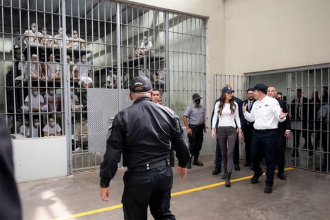 DHS Secretary Kristi Noem, El Salvador's Minister of Justice and Public Security Gustavo Villatoro, and a group of officials walk in front of a cell filled with men sitting on bunk beds.