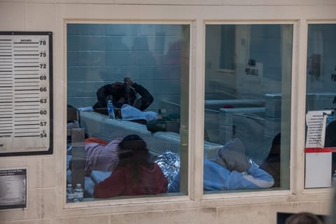 Interior shot of Ajo Border Patrol Station of a dirty, white concrete wall with three glass windows. A height chart and part of a blurry document containing the text: "etainee Suicide Risk arning Signs" hang on the wall. The glass windows provide a view into what appears to be a processing or holding cell, where multiple people are sitting or laying down in an open room.