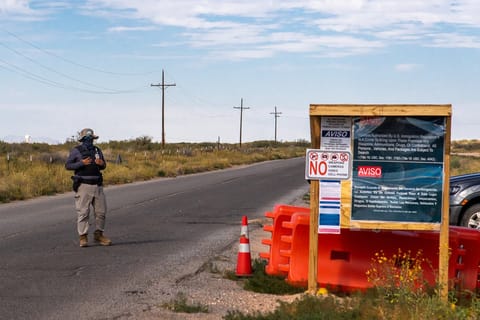 A masked guard stands in the road next to a sign with text: "NO weapons, cameras, video, cell phone" and other U.S. regulations.