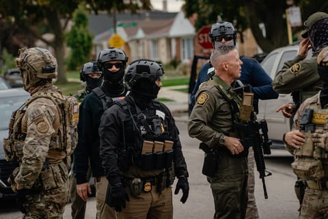 A group of federal immigration agents, including Chief Patrol Agent Gregory Bovino, stand together among parked cars on a street and survey what appears to be a residential area. Bovino holds a gun pointed at the ground and many of the agents wear helmets and camouflaged gear.