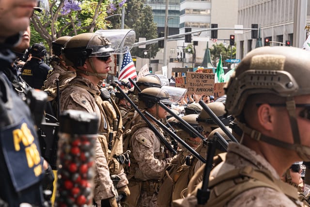 U.S. Marines and U.S. Soldiers wearing uniforms, helmets, and holding batons stand facing a crowd of protestors in Los Angeles, California.