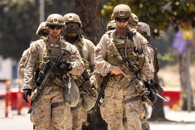A group of seven U.S. Marines wearing what appears to be combat gear and carrying guns walk in two lines down a city street.