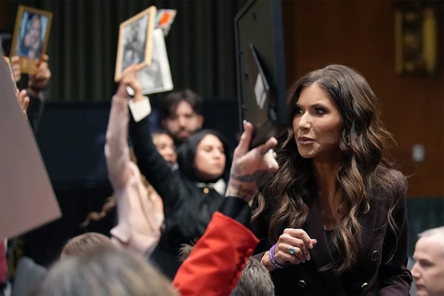 Protesters stand and hold photos as U.S. Secretary of Homeland Security Kristi Noem testifies before the Senate Judiciary Committee in the Dirksen Senate Office Building.