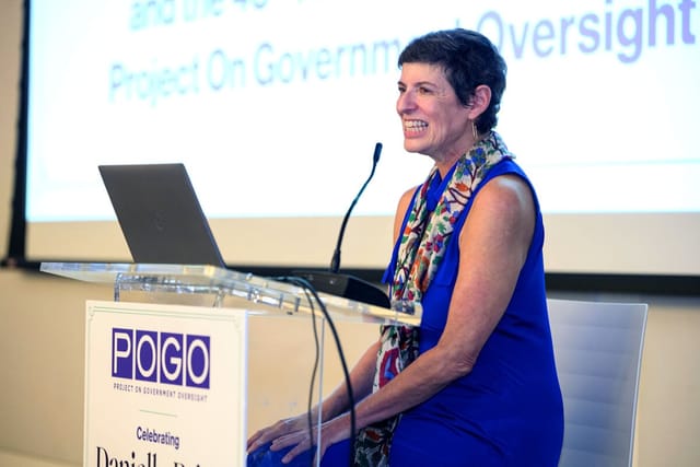 Danielle Brian speaks from a podium with the POGO logo. Behind her, a screen reads: Project On Government Oversight.
