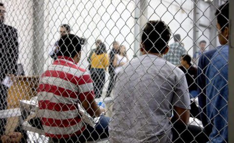 Detainees sit in a cell against a chain link fence.
